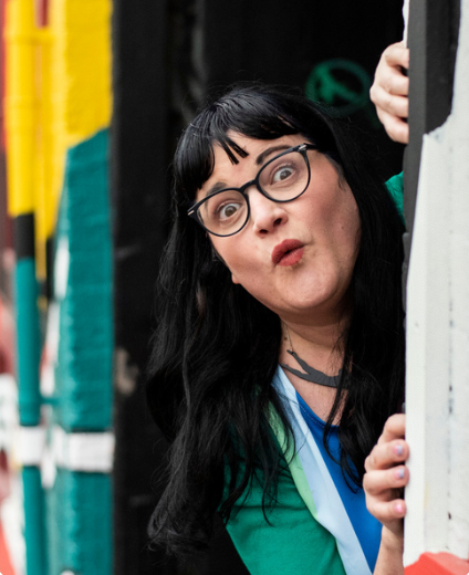 A picture of Heidi Mavir, a white woman with black hair and glasses, looking out from behind a curtain